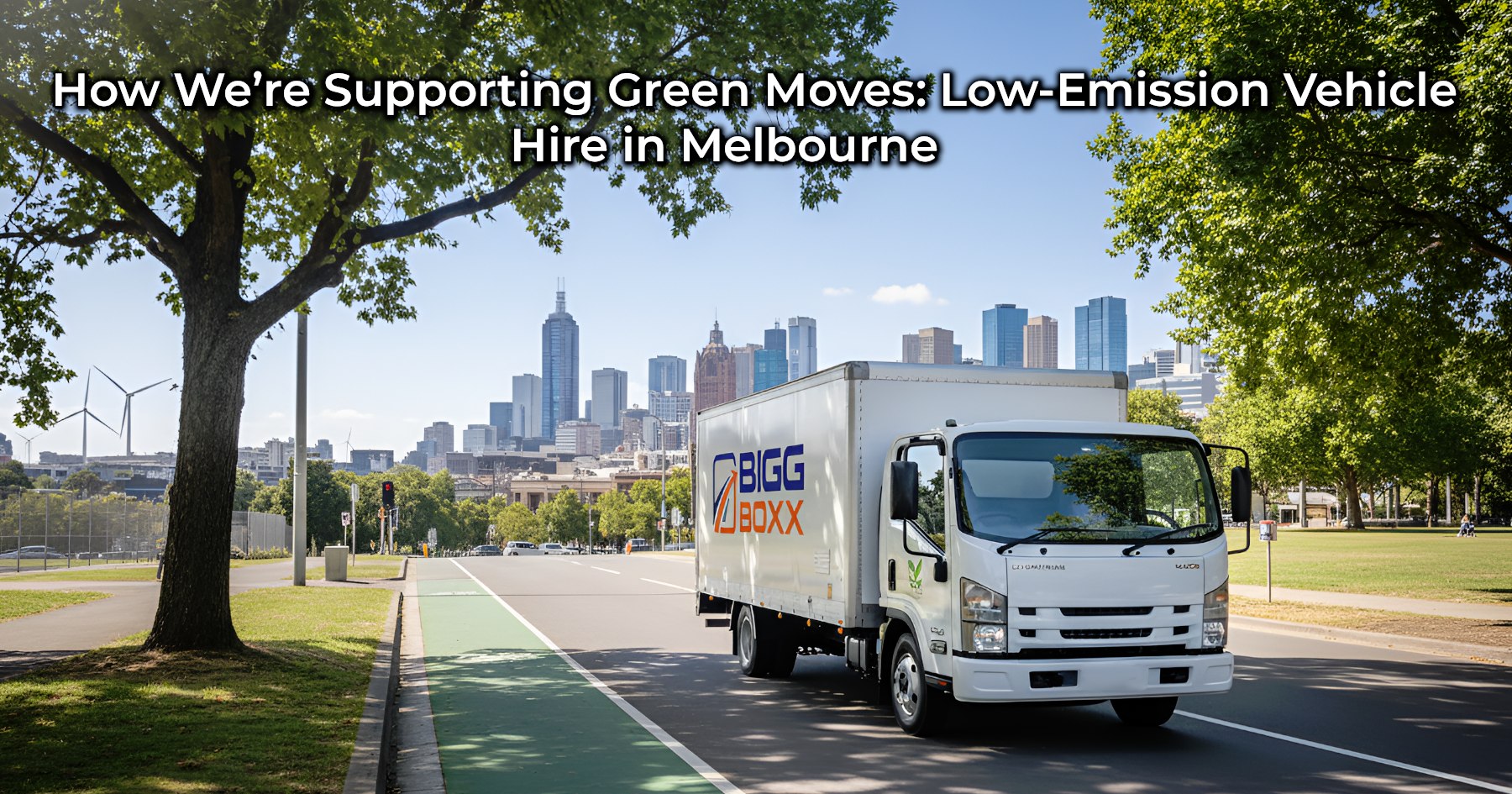 A white Bigg Boxx rental truck with the company logo on its side drives along a tree-lined road with a bicycle lane. In the background, a city skyline is visible under a clear blue sky | How We’re Supporting Green Moves: Low-Emission Vehicle Hire in Melbourne | Bigg Boxx Rentals