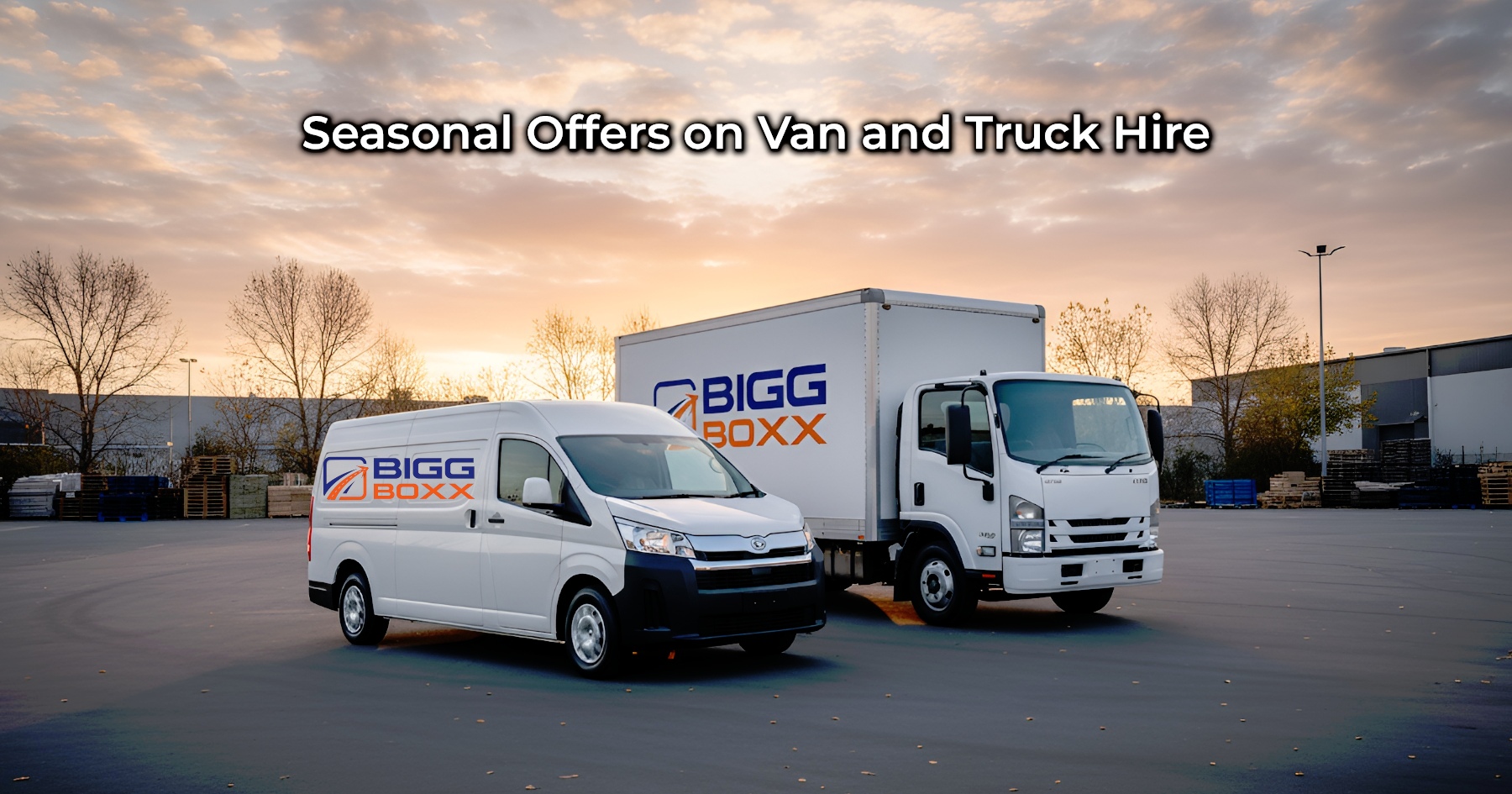 A white BIGG BOXX delivery van and a matching box truck with the company logo on their sides are parked side-by-side in a large asphalt lot at sunset, with industrial buildings and bare trees in the background | Seasonal Offers on Van and Truck Hire | Bigg Boxx Rentals