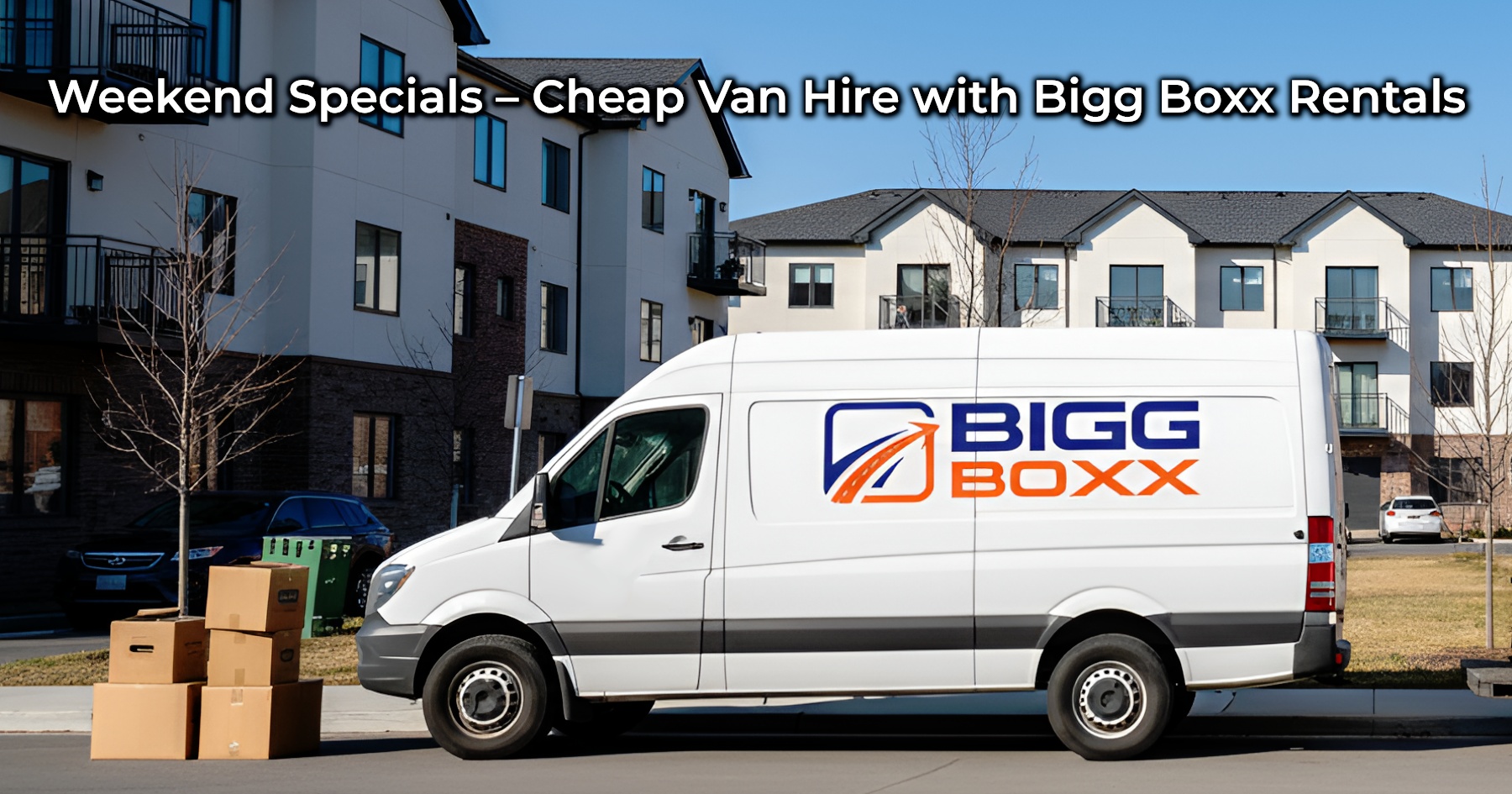 A white Bigg Boxx rental van, featuring the company logo on its side, is parked on a suburban street next to a stack of moving boxes. Modern apartment buildings are visible in the background under a clear sky | Weekend Specials – Cheap Van Hire with Bigg Boxx Rentals | Bigg Boxx Rentals
