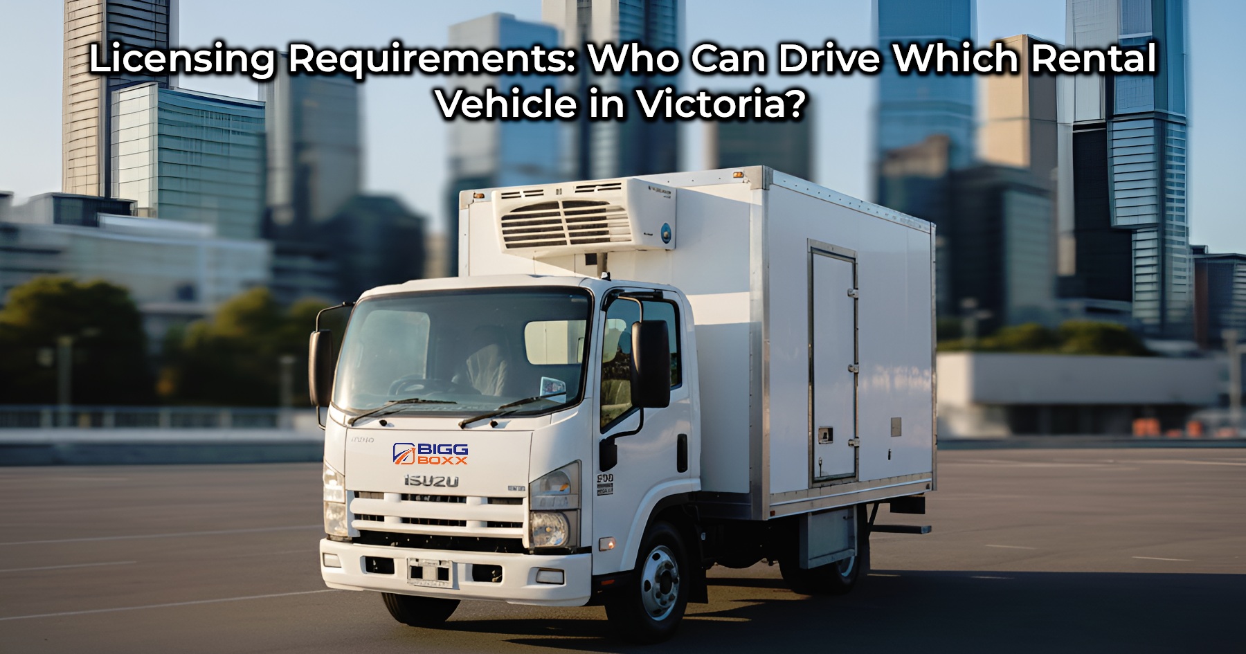 A white Isuzu refrigerated box truck is parked in a large, empty paved area with a modern city skyline of tall glass and steel buildings in the background under a clear, sunny sky | Licensing Requirements: Who Can Drive Which Rental Vehicle in Victoria? | Bigg Boxx Rentals